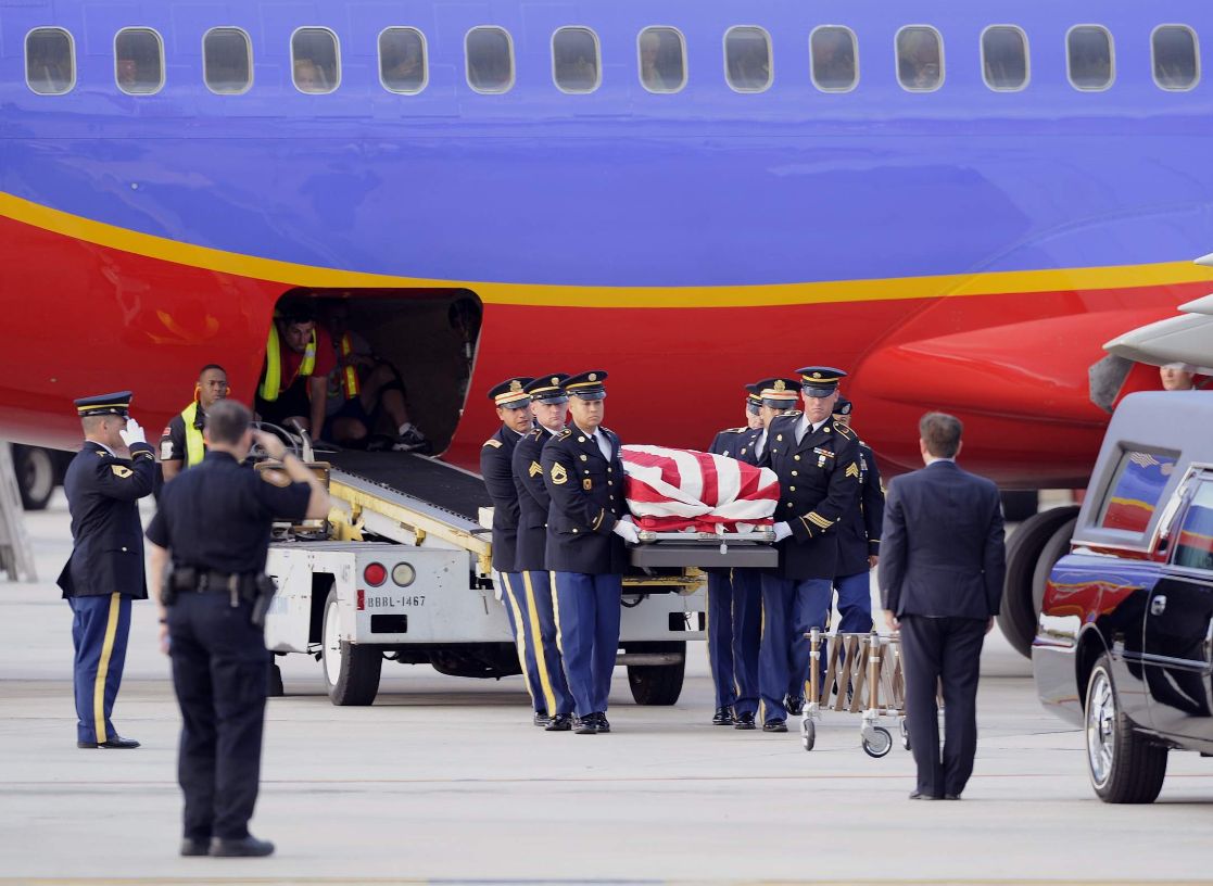 Soldiers carry a flag-draped casket from an airplane's cargo hold to a nearby hearse. The somber ceremony takes place on a tarmac, conveying respect.