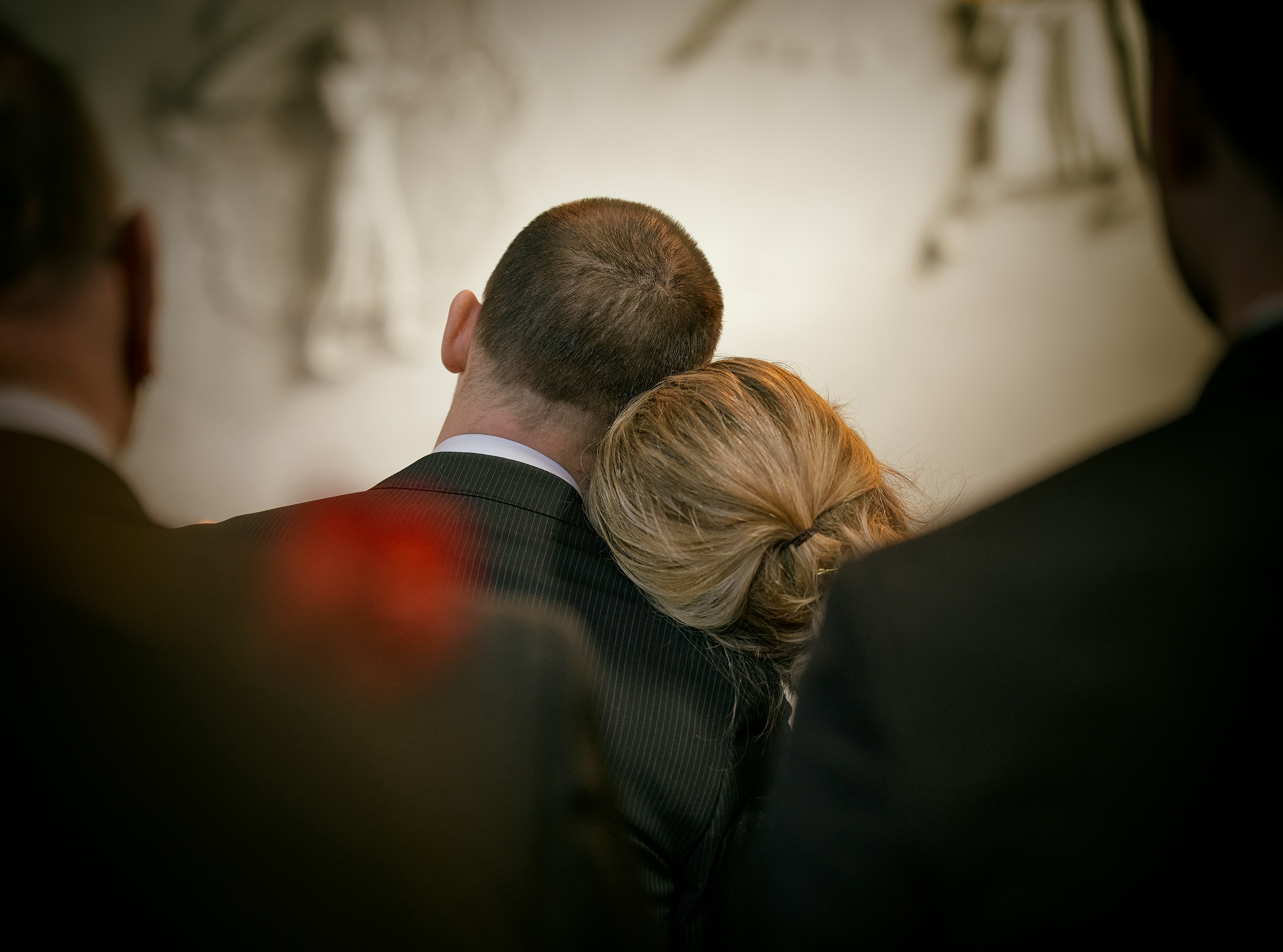 A couple in formal attire stands close, with the woman resting her head on the man's shoulder, creating a tender, intimate moment in a softly lit setting.