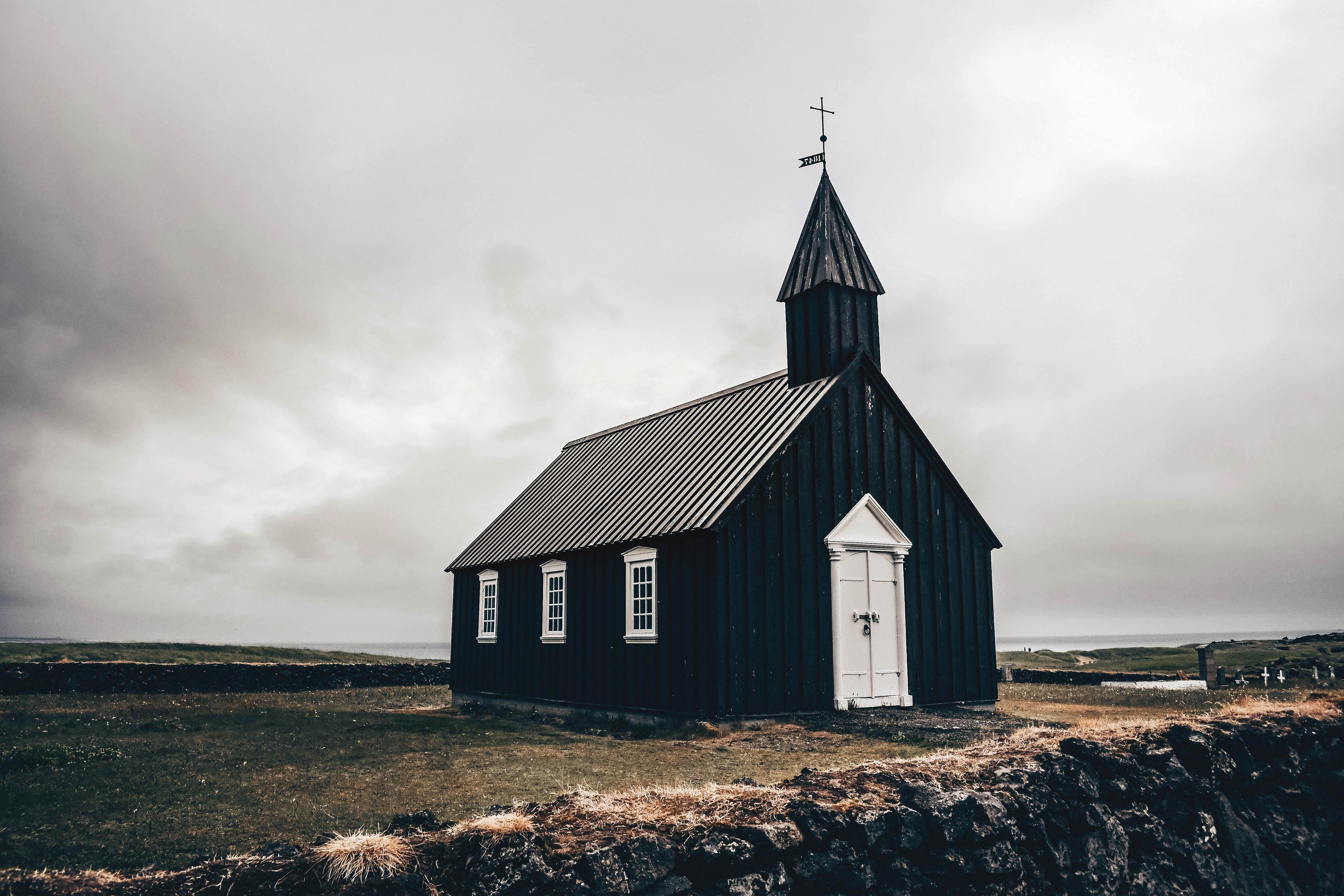 A small black wooden church stands solitary against a cloudy sky, evoking a serene yet dramatic atmosphere. The foreground features a grassy field and a low stone wall.