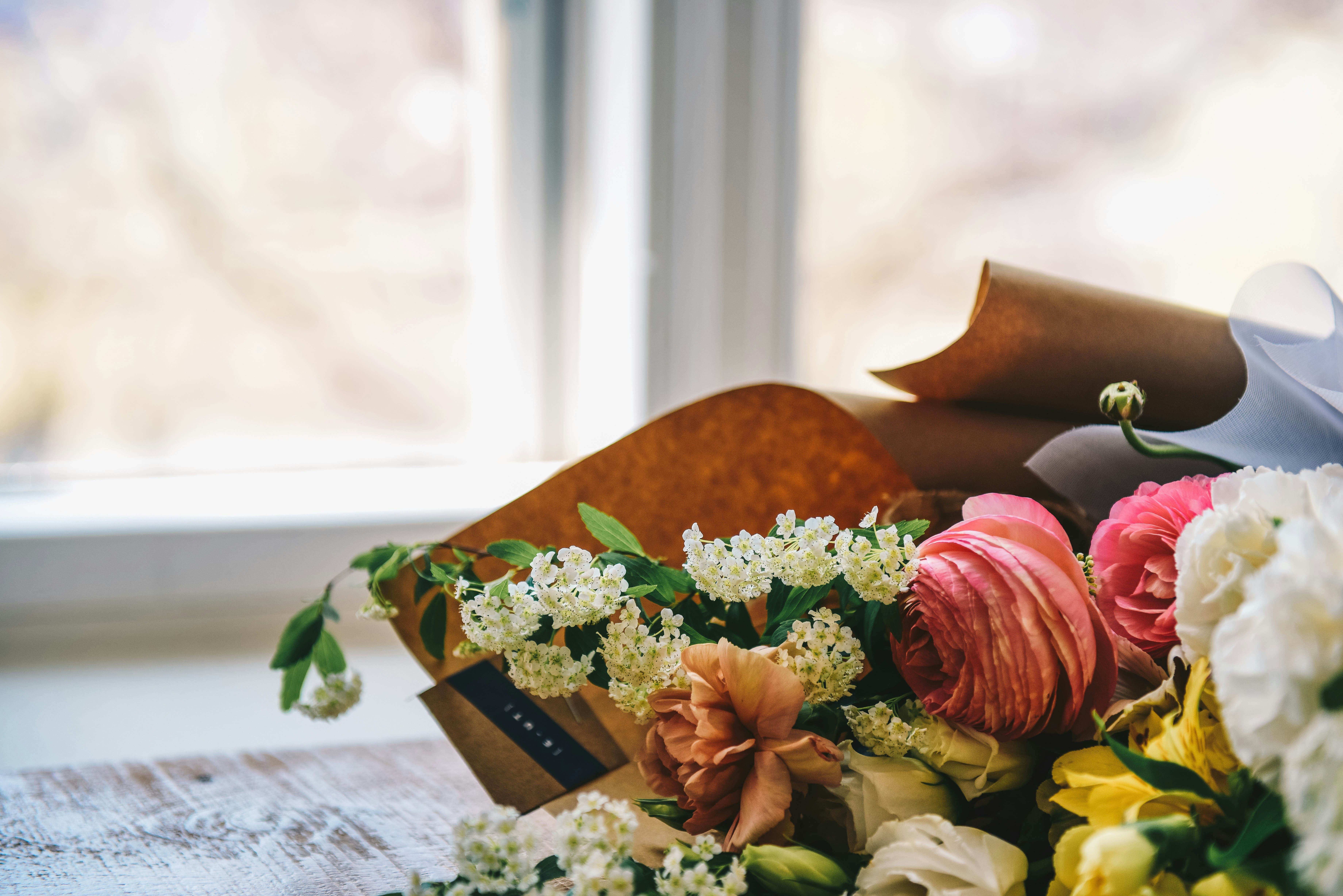 A vibrant bouquet of flowers, including pink and peach blooms surrounded by white blossoms and green leaves, rests on a rustic table near a bright window.